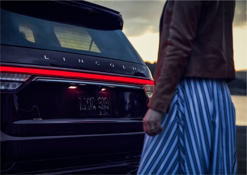 A person is shown near the rear of a 2023 Lincoln Aviator® SUV as the Lincoln Embrace illuminates the rear lights | Randy Marion Lincoln in Statesville NC