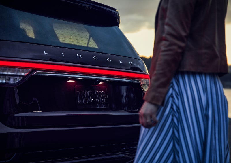A person is shown near the rear of a 2024 Lincoln Aviator® SUV as the Lincoln Embrace illuminates the rear lights | Randy Marion Lincoln in Statesville NC
