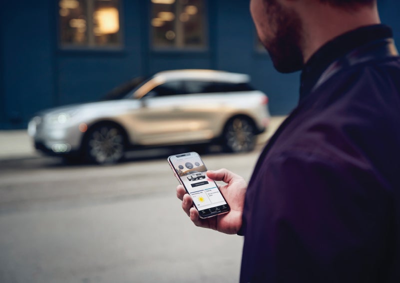 A man is holding his phone looking at the Lincoln Way™ App and we see his 2022 Lincoln Corsair SUV in the background against a blue brick wall | Randy Marion Lincoln in Statesville NC